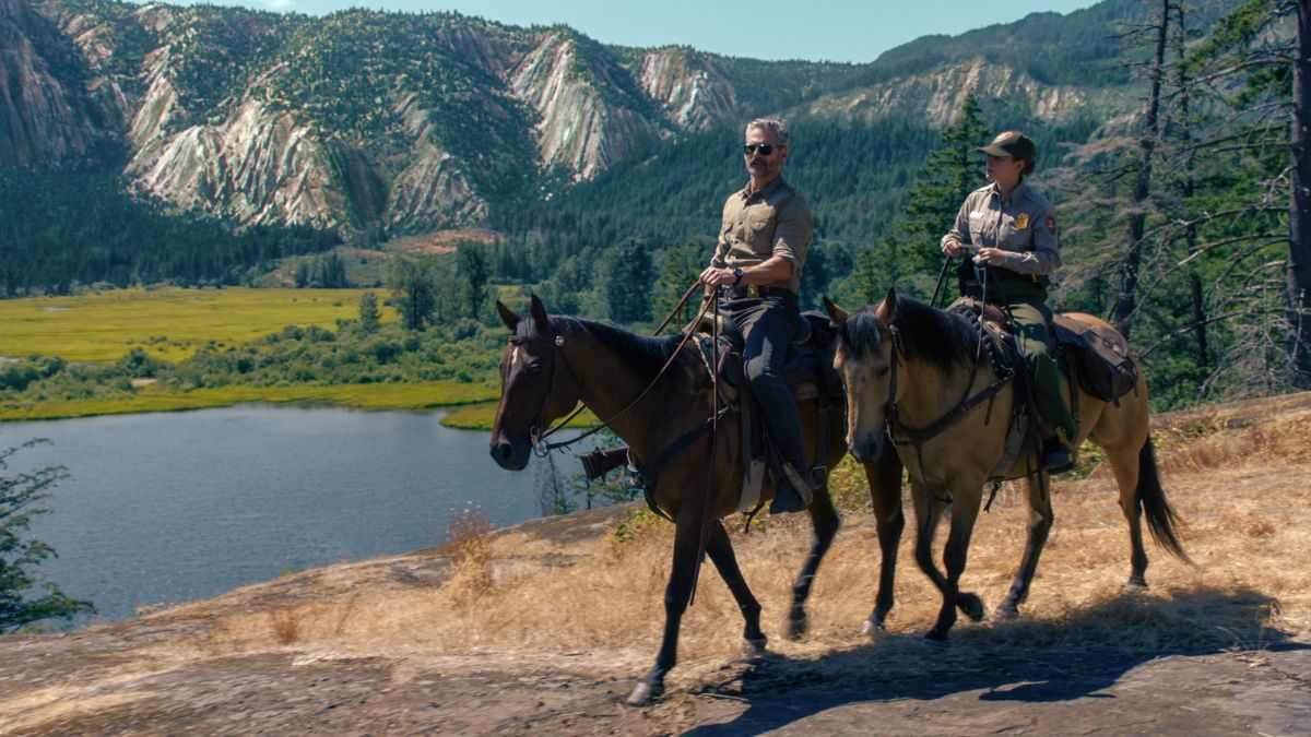 Kadr z serialu "Dzikość": Zdjęcie przedstawia dwoje jeźdźców konnych – mężczyznę i kobietę w mundurze służby parkowej – przemierzających słoneczny, skalisty teren. W tle rozciąga się malowniczy krajobraz z jeziorem, zielonymi łąkami, lasem i góram
